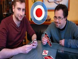 Ein Nutzer der Freizeitassistenz und sein Assistent sitzen an einem Tisch und spielen Karten. Sie lächeln und blicken in die Kamera.