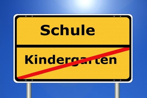 Bildbeschreibung: Gelbes Verkehrsschild bzw. Ortstafel mit schwarzer Schrift vor blauem Himmel und Sonnenschein im Hintergrund. Das Schild ist durch eine schwarze Linie in 2 gleich große Bereiche geteilt. Im oberen Bereich der Verkehrstafel steht das Wort „Schule“ geschrieben, im unteren Bereich das Wort „Kindergarten“. Das Wort „Kindergarten“ ist mit einer roten Linie durchgestrichen, die von links unten nach rechts oben diagonal verläuft.