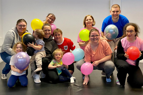 Das Bild zeigt zeigt lächelnde Personen in unterschiedlichem Alter, auch Kinder sind dabei. Alle haben einen Luftballon in der Hand und sitzen, knien oder hocken am Boden.