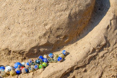 Mehrere kleine, bunte Glaskugeln liegen dicht beieinander in einer schmalen Rinne aus Sand. Das Bild steht sinnbildlich für Vielfalt, Inklusion und Gemeinschaft und begleitet die Sammlung der Sommerferien- und Urlaubsangebote 2026 von Integration Wien.