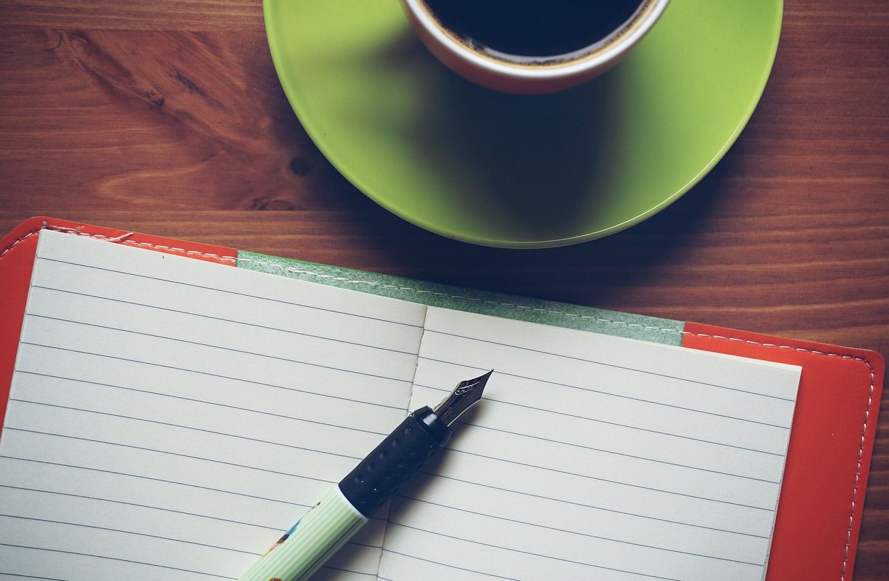 Notizbuch auf dem Füllfeder liegt. Vor dem Notizbuch steht eine Tasse befüllt mit Kaffee.